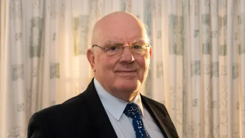 Sue Jones Howard Parkin, a bald man, with glasses, wears a suit and astronomy themed tie