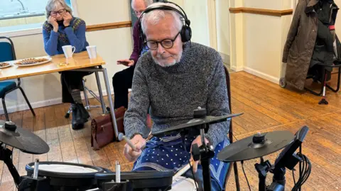 BBC Nick Foster is sitting at an electric drum kit. He is wearing a grey jumper and has headphones on.