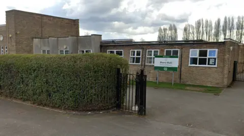 A low brick building behind a hedge with a fence in it. A sign outside the building reads "Perry Hall Primary School".