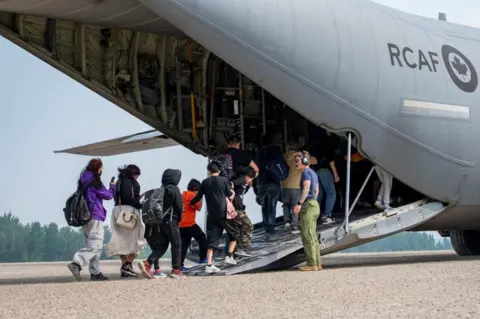 Canadian Armed Forces official assists residents onto a transport aircraft during a wildfire evacuation