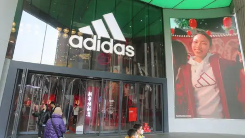 Adidas shopfront. People are walking through the front doors of a big glass shopfront with the adidas logo above it. The logo is three left-facing rectangles at 45 degrees increasing in size from left to right. On the right of the shop is a massive banner of a model happily wearing an Adidas hooded top. She is smiling broadly.