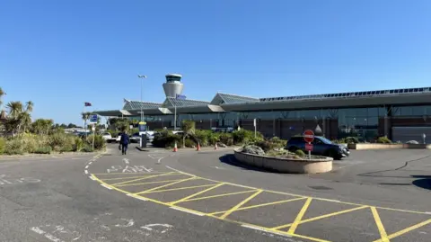 Jersey Airport on a sunny and clear blue sky day. There is a man walking up to the airport building which is largely glass with three triangular shaped glass roofs. The air traffic control tower, a silver spherical building, can be seen just past the airport. 