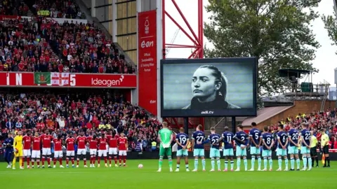 PA Media Two rows of football players with their arms around each other with a black and white picture of Maddy Cusack on a large screen at a full stadium
