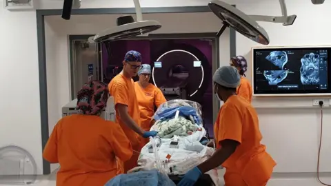 Supplied Medical teams in orange scrubs wheeling a small child into an operating theatre
