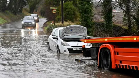 Pacemaker Cars on a flooded road near Drumbo. A white Renault is stranded in the water with its bonnet open. A red recovery vehicle is partially visible, and two other cars are travelling towards the water. 
