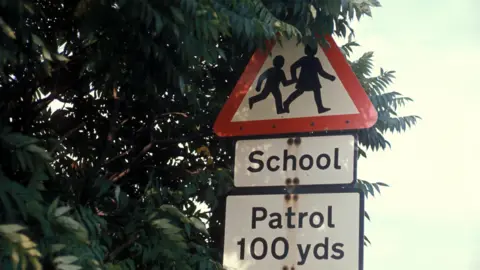 A triangular road sign signifying a school next to a tree that is almost covering the top of the sign. The lower sign warns drivers that there is a crossing patrol 100 yards ahead.