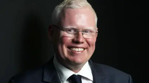 ABC News/Justin Huntsdale Gareth Ward, wearing a pair of glasses, looks at the camera for a portrait style shot with a black background behind him
