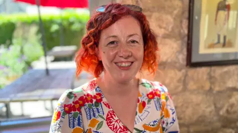 A woman with short, curly red hair is sitting indoors by a window, facing the camera. She wears a colourful patterned top, and sunglasses rest on top of the head. Behind, a stone wall and a framed picture are visible, while outside the window a red umbrella and greenery can be seen in soft focus.
