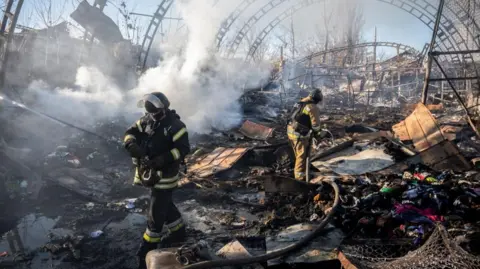 Firefighters walking through smoke and debris at the site of where a Russian drone has hit, with large oval metal structures visible in the background.