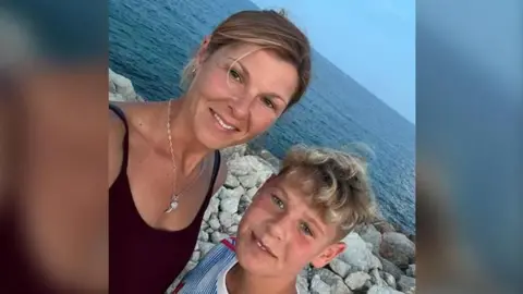 A woman in a black dress with her hair tied up standing next to a young, blonde boy in a blue and white Manchester United shirt. They are standing in front of the ocean.