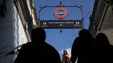 Getty Images Silhouette of commuters walking up the stairs of a London Underground subway station entrance, framed by the Westminster Station sign and iconic red and blue roundel against a clear blue sky.
