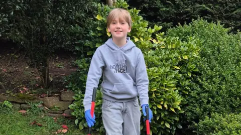Young boy standing in a garden, smiling at the camera while holding two litter pickers, wearing a grey hoodie, blue gloves and casual trousers, with green bushes and trees behind him