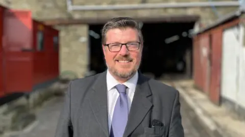 Andie Cowie, short dark hair, a short beard and glasses. Wearing a dark grey suit with a lilac tie.