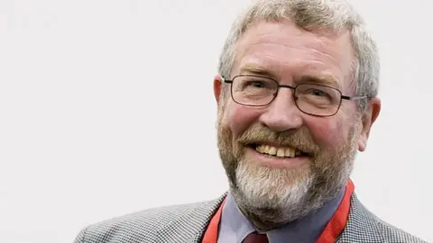 Supporters Direct Brian Lomax in a suit and tie, smiling at the camera. He has a beard and short grey hair and is wearing glasses.