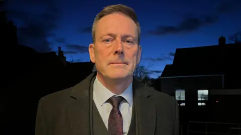 Tony Fisher/BBC Nick Bell, standing outside in the dark, with houses behind him, and a dark sky. He is looking straight at the camera, with short hair, wearing a suit, shirt, tie, and coat. He looks serious. 