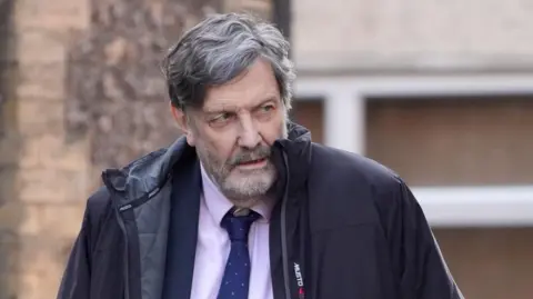 Alan Harris walking. He is wearing a pink top with a blue tie and blazer. He is wearing a black coat. The background shows a brown wall