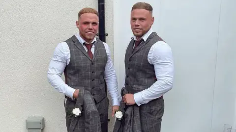 Corey and Liam dressed in white shirts and grey waistcoat and trousers with maroon tie. They both have short light brown hair and a short beard and are carrying grey jackets with white carnations in the buttonhole.