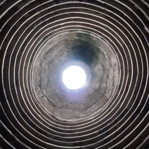Charlie Hood An upward view inside a circular stone structure, with concentric rings lining the interior walls and a bright opening of daylight visible at the top, creating a tunnel-like effect.