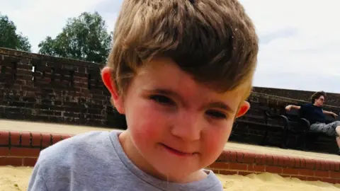 Jack Moate in front of a brick sea wall on a sandy beach. He has brown hair and is wearing a grey sweater.