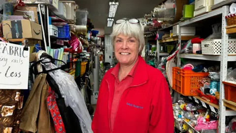 A woman in a red zip up jumper that says 'Saltash Scrapstore' and a red top. She has short hair and glasses on her head. She is smiling at the camera. Around her is various items of scrap on shelves.