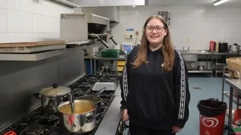 Rachel Fairless is standing smiling in the kitchen beside a big pot on a large hob. She is wearing a black top, glasses and has long fair hair. 