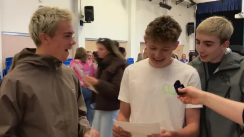 Three teenage boys stand in a busy hall reading exam results while someone holds a small microphone toward them, with others talking and blue chairs lined along the walls in the background.
