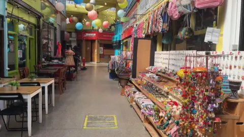 A corridor of shops in an indoor mall. There are tables and chairs outside a cafe on the left and a display of goods outside a shop on the right. 
