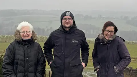 Three people, Kate Culverhouse of the Banks Group, Andrew Wadds of Durham Wildlife Trust and Louise Harrison of the Banks Group, standing in a line in front of a field. It is a grey day and the hills behind them are covered in mist. Kate has white, short hair and is smiling into the camera with one eye closed. She is wearing a big, dark coat and has round glasses on. Andrew is squinting at the camera and is wearing a beanie. He has his hands in his pockets and has a coat on with the Durham Wildlife Trust logo. Louise has dark hair and is wearing glasses. She is wearing a dark coat with her hands in her pocket and is smiling.