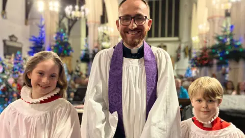 Jon Wright/BBC A vicar stands with two choristers in a church. There are Christmas trees in the background.
