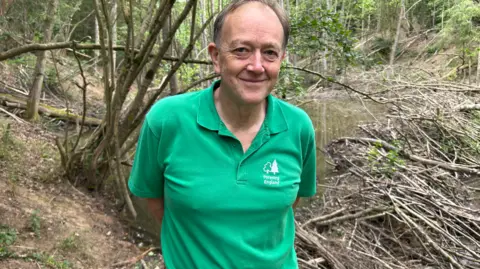 A man with short grey hair is standing in front of a number of trees in a forest. He is wearing a green jacket with a tree logo and "Forestry England" emblazoned at the front.