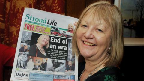 Jo Barber with blonde hair holding up a newspaper which has a headline and images of her. She was given the newspaper when she retired from the paper. 