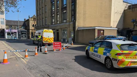 Alex Moss/BBC Police car parked in front of a closed off street, which is being guarded by a police officer behind tape.