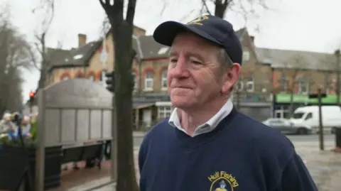 BBC A man is smiling and looking to the side, wearing a navy cap with a yellow logo and a navy jumper, with a yellow logo that says 'Hull Fishing' at the top, with a white shirt underneath. The background is blurred but you can see the corner of a street with cars parked, and to the left people can be seen congregating behind a placard. 