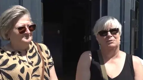 LDRS Two women with silver hair are wearing sunglasses; one wears a beige and cream patterned top and the other wears a black top. There is a black door behind them.