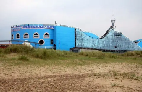 Geograph/Steven Daniels The seafront-facing side of the scenic roller coaster, which is painted blue and the side undergoing repainting. There is grassy and sandy land in the foreground.