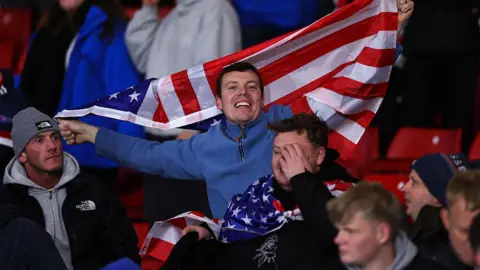 Getty Images A person in a stadium holds a large United States flag spread out behind them. Other fans are seated nearby, some also draped in American flags. The background shows red stadium seats and people wearing winter clothing.