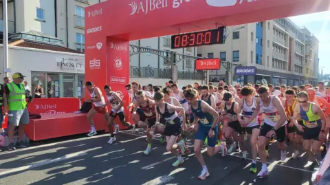 A group of runners is crossing the start line in a city centre with houses on one side of the road.
