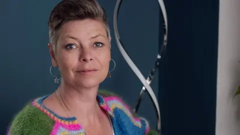 Laura Marfell-Williams, with swept back short hair and hooped earrings. She is wearing a striped green, bright pink, grey, blue and yellow woollen cardigan on top of a white vest top, and is sitting against an inky blue wall.