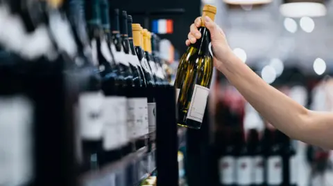 A woman grabs a bottle of wine from a shop shelf.