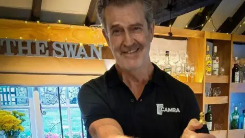 CAMRA Hollywood film star Rupert Everett wearing a black top with the CAMRA logo on it standing behind the bar in the Swan pub pulling a pint. In the background there is a 'The Swan' sign, glasses and bottles, fairy lights and a vase of yellow flowers. 