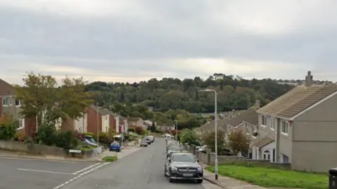 A residential road. Terraced houses line both sides of the street. Cars are parked on the road in front of the houses. There are trees in the distance. A green patch of grass is in front of one house at the end of the street.