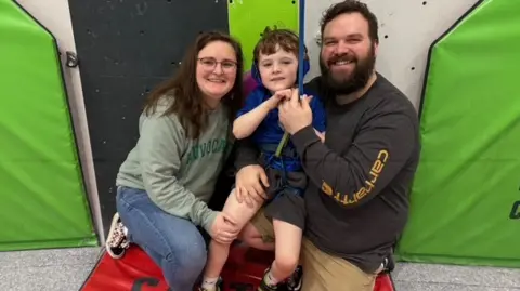 A young boy in a blue top and blue headphones attached to a climbing rope. His mum is on the left of him and is wearing a grey jumper and blue jeans. She is wearing glasses. His dad is on the right and is wearing a dark grey jumper with beige trousers. He has a dark brown beard. The three of them are hugging and smiling, sat against a green climbing wall. 