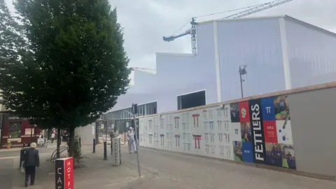 Photograph of the new Fettlers shopping district being built in Wigan town centre. The image shows wooden boarding in front of the indoor market building.