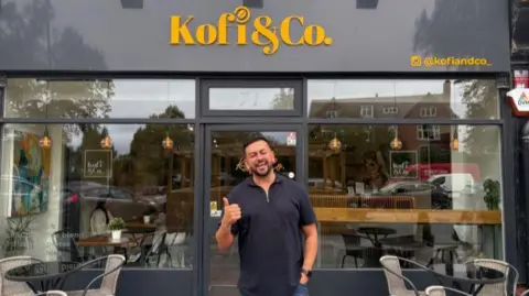 Louis Reynolds/BBC Harry Limbert stands doing a thumbs up gesture in front of a cafe called Kofi & Co