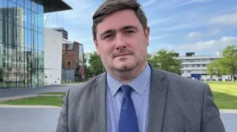 Chris Cooke standing next a building with glass front and grass behind him. He is wearing a grey jacket, blue tie and blue checked shirt.