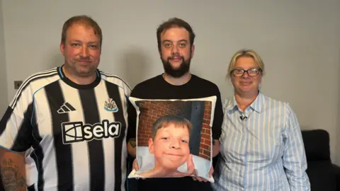 Craig Anderson, who is wearing a black and white Newcastle United football shirt, stands next to Aaron's brother, Lewis, who is wearing a plain black shirt and is holding up a cushion. The cushion has a picture of Aaron on it, which shows him smiling and giving a thumbs up. On the other side of Lewis is Aaron's mother Donna, who is wearing a blue and white striped T-shirt.