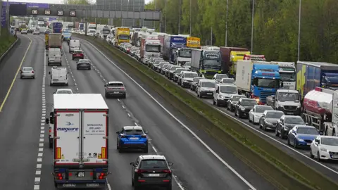 A very busy main road. There is a traffic jam on the road going in the direction toward the camera. On the side of the road going away from the camera, traffic is minimal. In the distance there is a sign which says 'M50 TOLL'. 