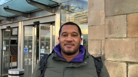 Simon Coombs stood outside Sheffield train station. He has short, dark hair and is wearing a green jacket with a purple sweater underneath.