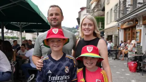 A family pose and smile at the camera. Dan and Christina, stand at the back of the group while their children, Dylan and Mali, stand behind them. Dan is wearing a grey top and has his hand on Dylan's shoulder. Christina is wearing a black top and stands behind Mali. Dylan is wearing a blue sports top while Mali is wearing a red and yellow top and hat.