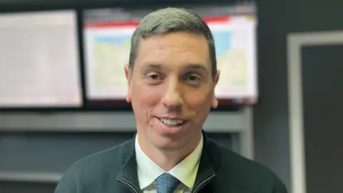 A man with short hair staring at the camera. He is wearing a shirt and tie under a jumper. 
